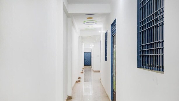 A long, narrow hallway with white walls, a tiled floor, and a blue window with bars. - Hotel kartikey (Phulpur)