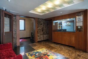 Um saguão de hotel com balcão de recepção, uma cadeira vermelha e piso de mármore.