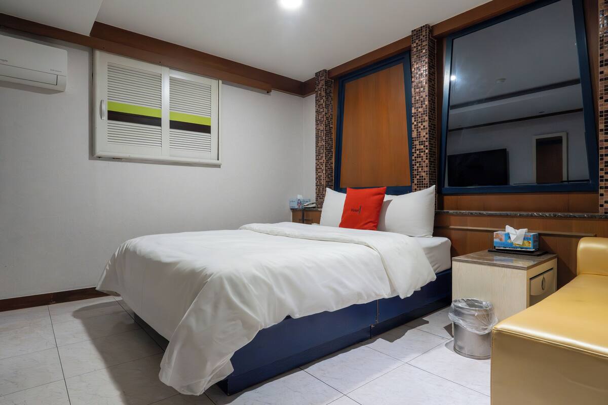 A hotel room with a bed, a bedside table, a trash bin, a small nightstand, and a window with curtains.