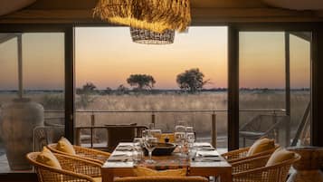 A dining area with a table set for six, overlooking a savannah landscape at sunset.