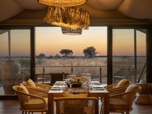 A dining area with a table set for six, overlooking a savannah landscape at sunset. - Thitaka River Lodge by NEWMARK - All Inclusive (Kongola)