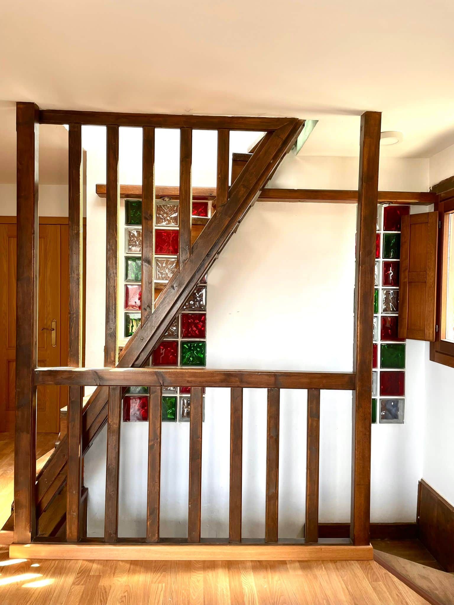 A wooden staircase with a glass railing, leading to a room with a white wall and a shelf with colorful glass bottles.