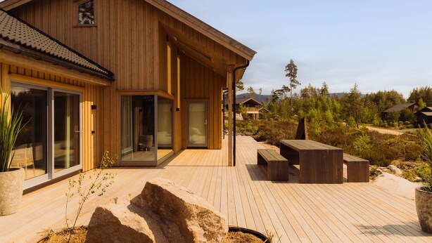Une maison en bois dotée d’une terrasse, de grandes fenêtres et offrant une vue sur le paysage environnant.