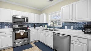 Uma cozinha moderna com armários brancos, micro-ondas, forno e lava-louças de aço inoxidável, um backsplash de azulejos azuis e uma janela com persianas.