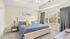 Quarto com cama, mesa de cabeceira, abajur, janela com cortinas e ventilador de teto.