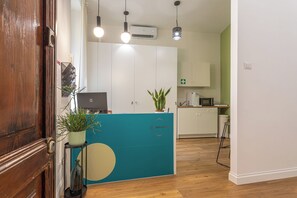 Un área de recepción con puerta de madera, mostrador verde azulado, plantas en macetas, una computadora portátil y un armario blanco.