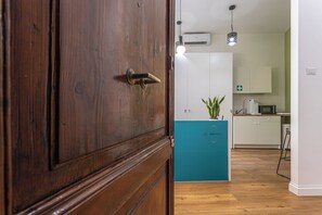 Una puerta de madera con manija de latón, una encimera color verde azulado con una planta, y una zona de cocina con armarios blancos y un microondas.