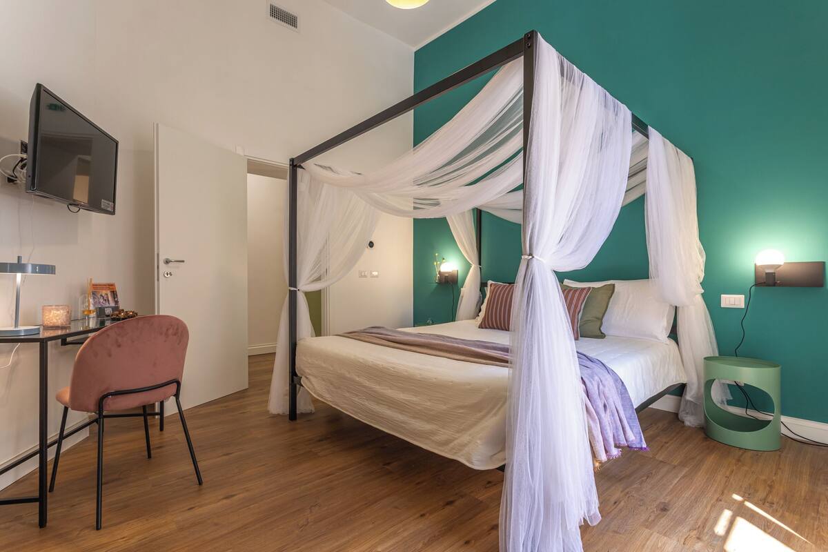 Una cama con dosel con cortinas traslúcidas, un escritorio con silla, un televisor y una mesita de noche verde.