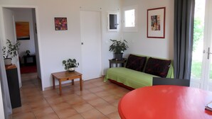 A room with a green sofa, a red table, a small wooden table with a plant, and a door leading to another room.