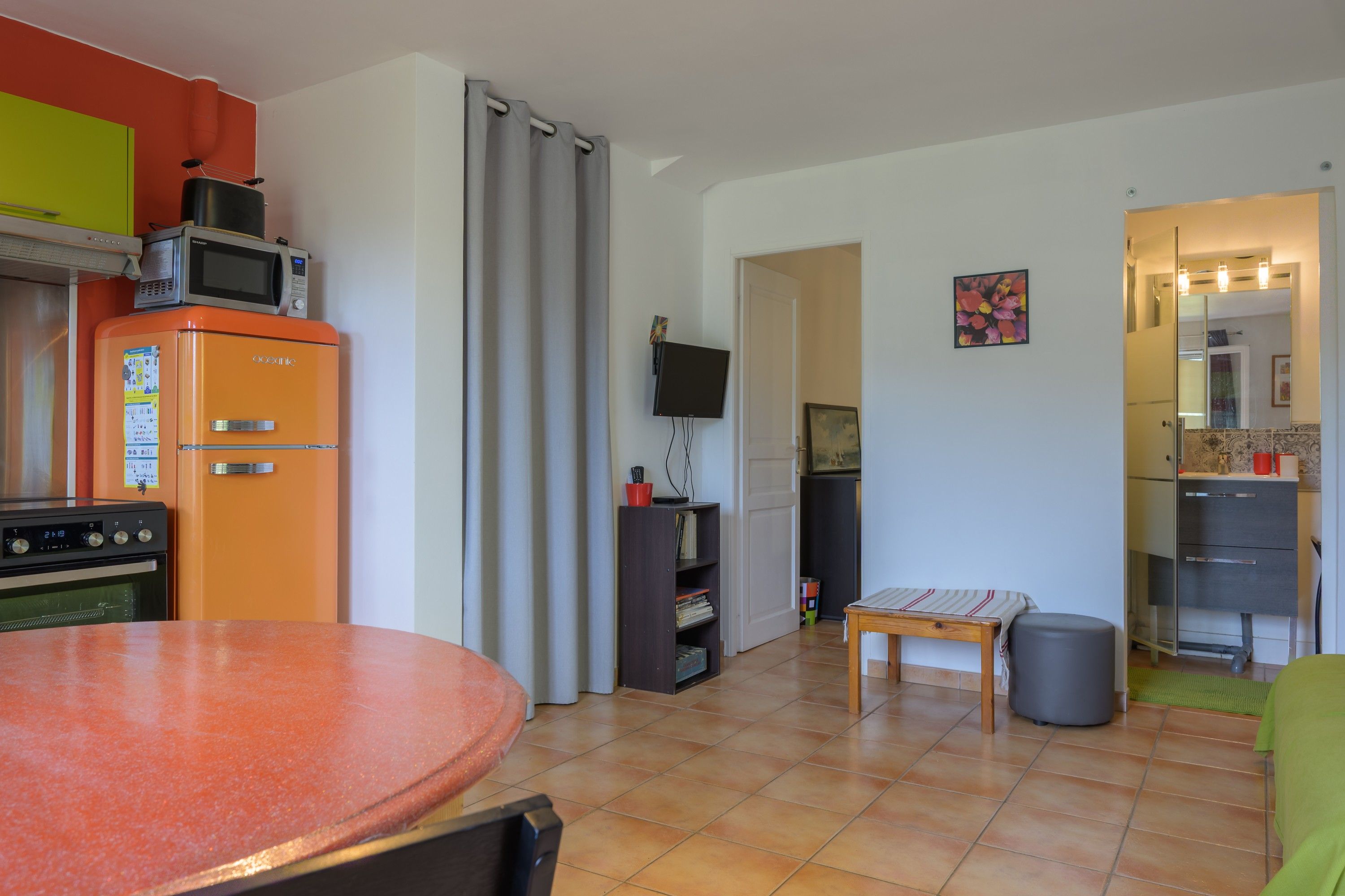 A compact kitchen with a refrigerator, oven, and microwave. There is a dining table with chairs, a small table with a single chair, and a television mounted on the wall.