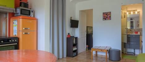 A compact kitchen with a refrigerator, oven, and microwave. There is a dining table with chairs, a small table with a single chair, and a television mounted on the wall.