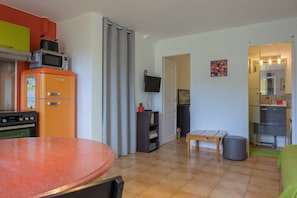 A compact kitchen with a refrigerator, oven, and microwave. There is a dining table with chairs, a small table with a single chair, and a television mounted on the wall. - Gîte de Maric: Among Olive Trees with Pool, Stunning Mountain Views (Aubres)