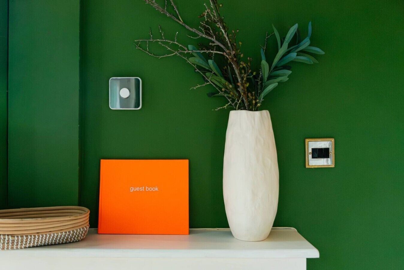 Un mostrador de recepción con un libro de visitas, un jarrón con ramas y un interruptor de luz en la pared.
