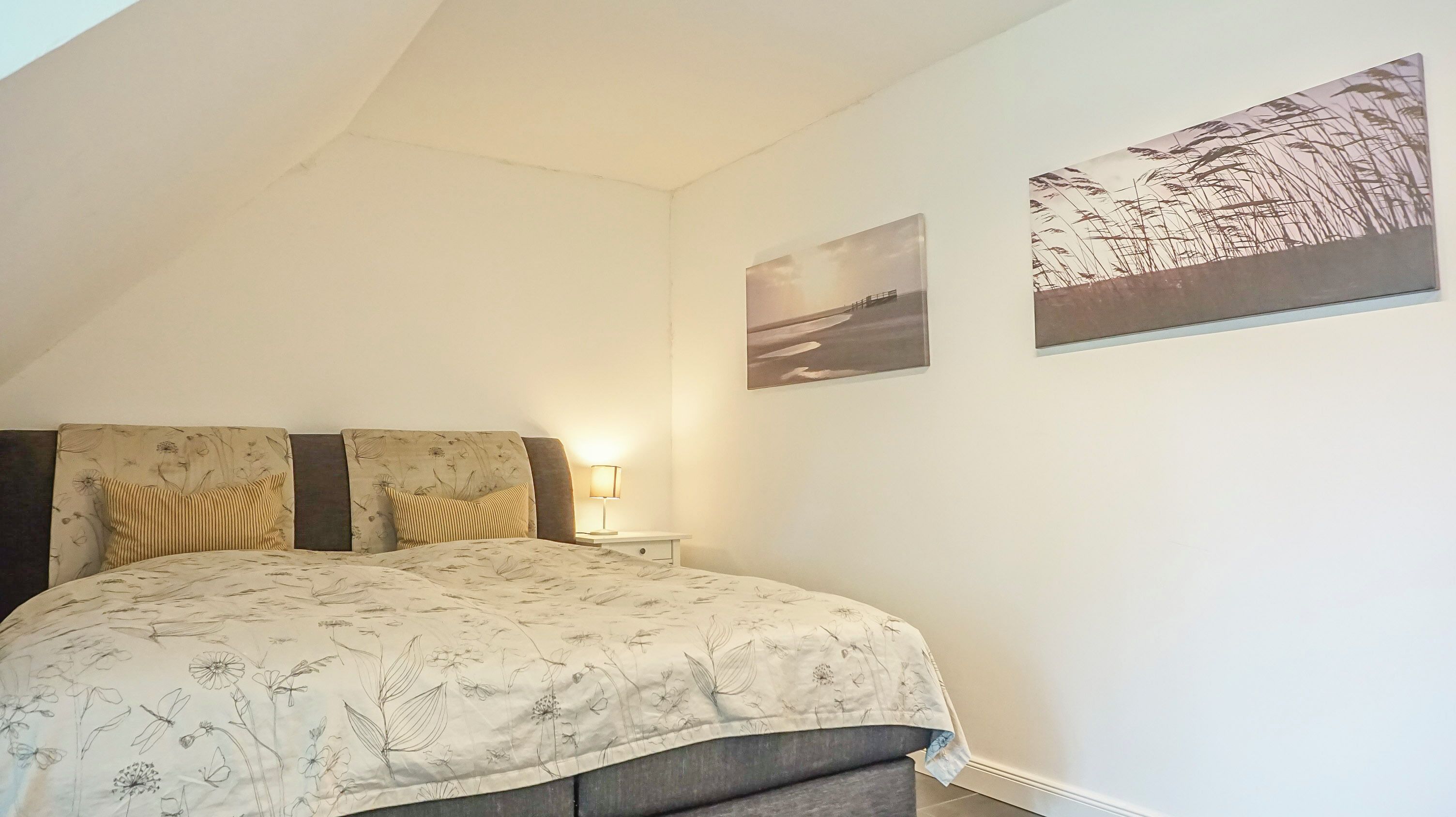 Une chambre à coucher avec un lit, deux oreillers, une table de chevet, une lampe et deux tableaux accrochés au mur.