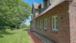 A brick building with a sloped roof, a pathway leading to the entrance, and a tree-lined background. - Warftkroog Thatched-Roof House: Thatched-roof house for 20 people with a sauna and hot tub (Katharinenheerd)