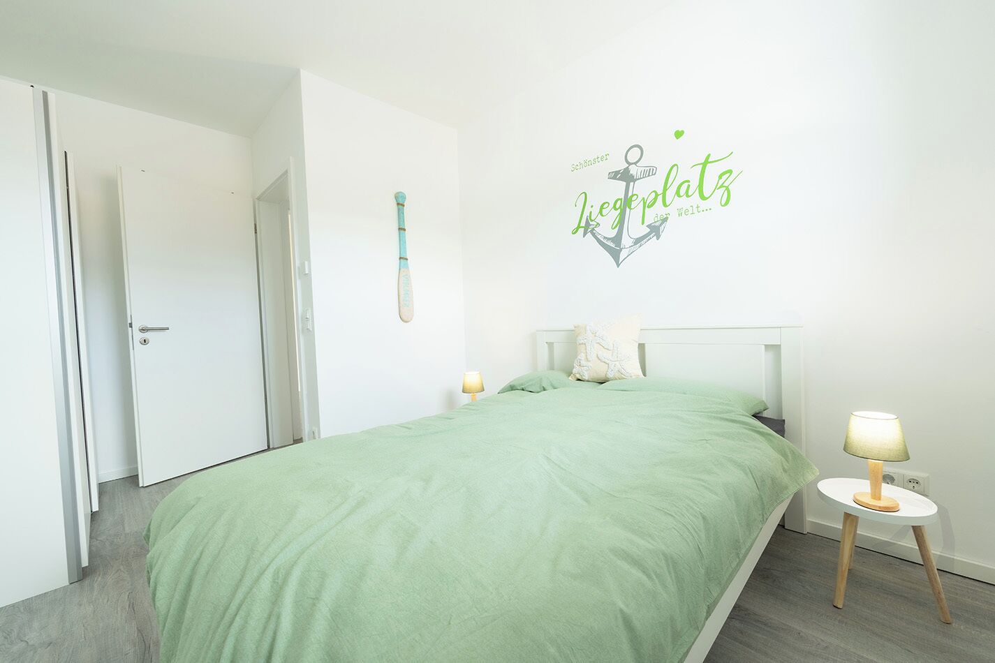 A bedroom with a green bedspread, a white headboard, a wall decoration, a lamp on a bedside table, and a door.