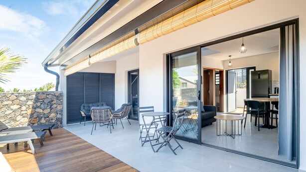 A modern outdoor patio with a stone wall, wooden deck, and glass doors leading to an interior dining area. - House with AC and WiFi - 2 guests, near St-Pierre (Saint-Pierre)