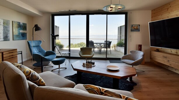 A modern living room with a sofa, chairs, a coffee table, and a TV. There is a balcony with a view of the sea. - Modern duplex, panoramic view - 6 guests (Évian-les-Bains)