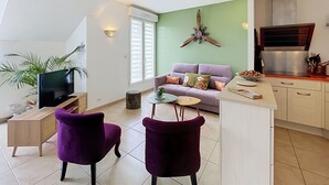 Ein modernes Wohnzimmer mit einer Couch, zwei violetten Sessel, einem Fernseher, einer Pflanze und einer Küchenzeile mit Herd und Schränken.