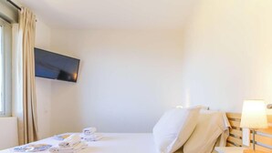 A hotel room with a bed, a TV mounted on the wall, a bedside table with a lamp, and a window with curtains. - Apartment, pets welcome, sea view - 4 guests (Le Lavandou)
