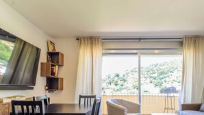 A living room with a dining table, chairs, a sofa, a TV mounted on the wall, and a balcony with a view of a hillside. - Apartment, pets welcome, sea view - 4 guests (Le Lavandou)