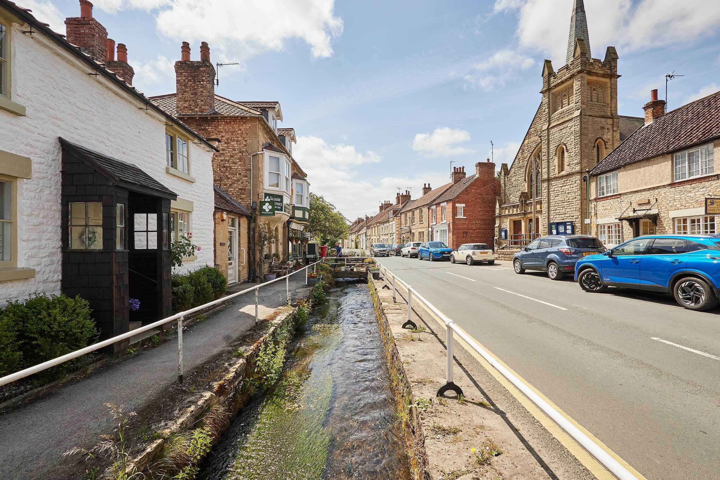 A street with buildings, a narrow water channel, and parked cars.