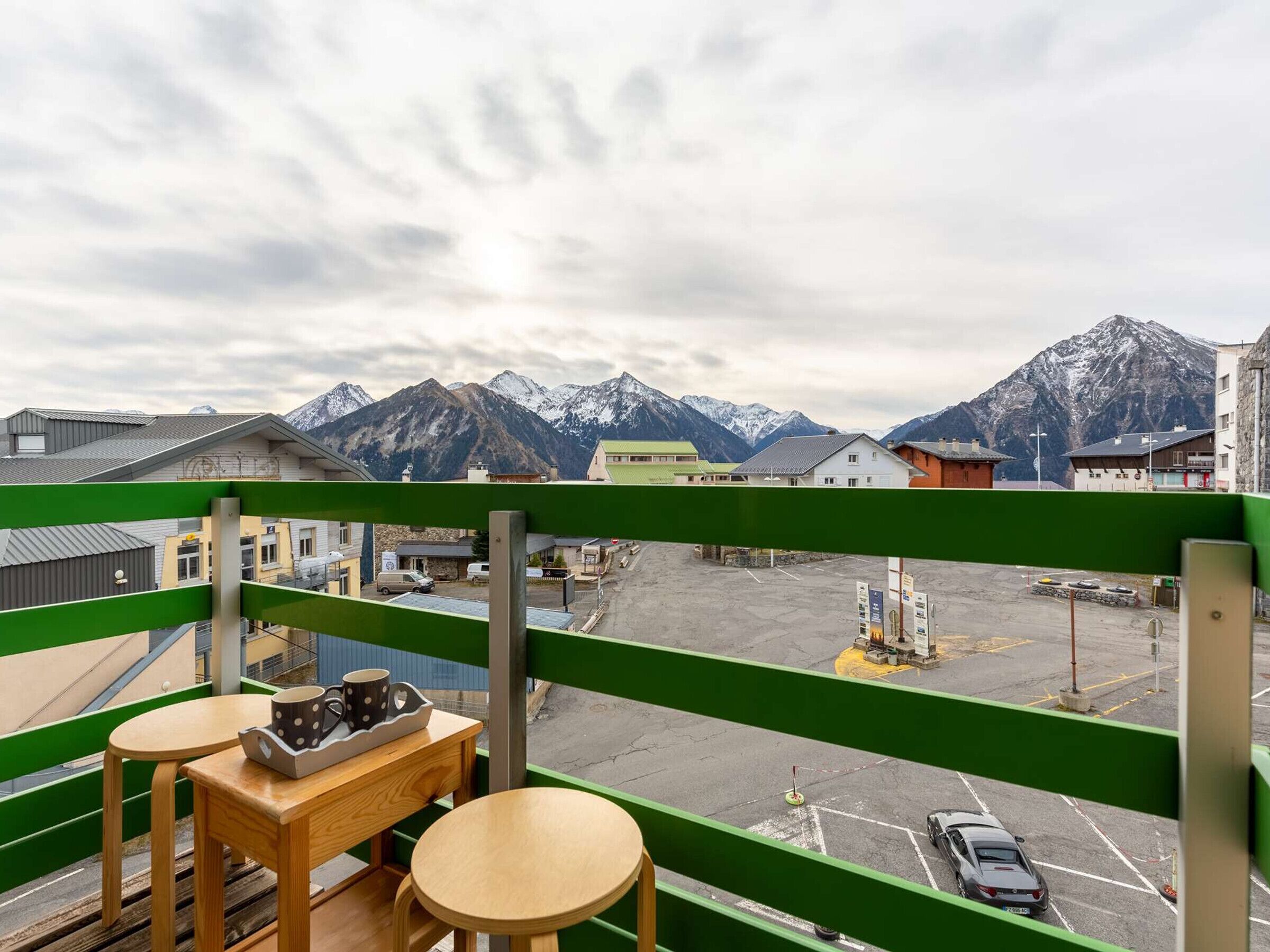 A balcony with a view of snow-capped mountains, a parking area, and a table with a mug.