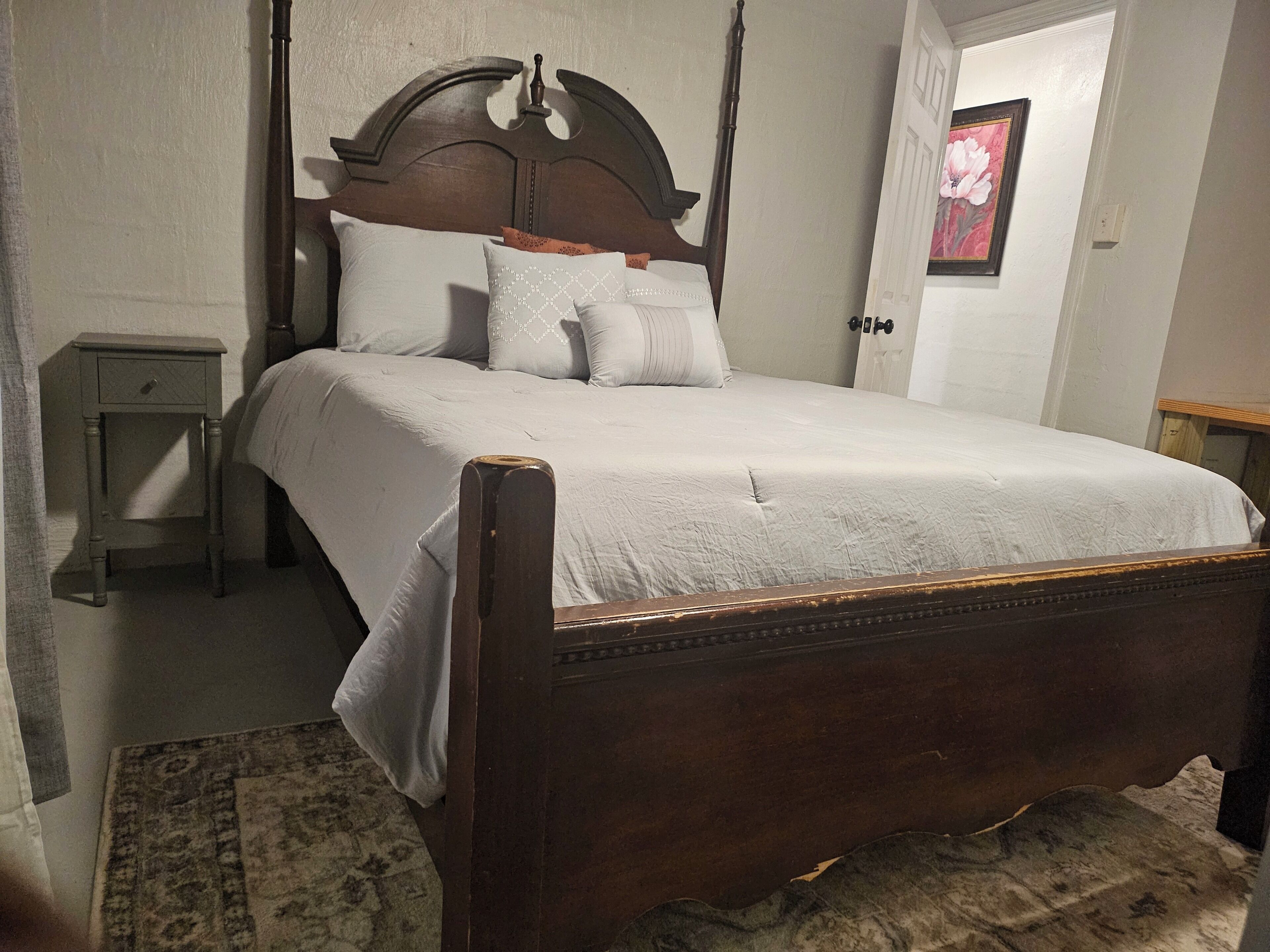 A bedroom with a wooden four-poster bed, a nightstand, a rug, and a framed picture on the wall.