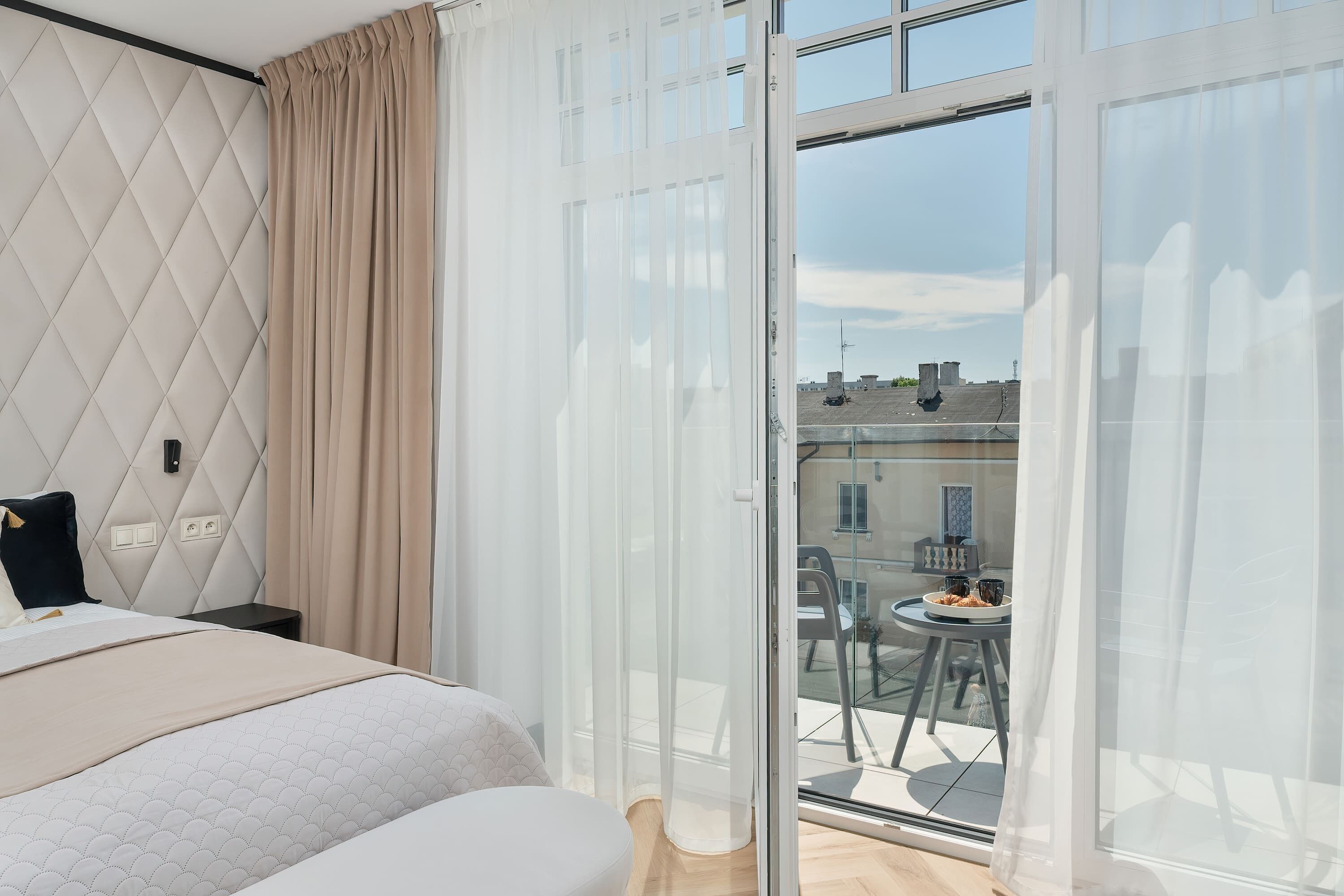 Moderni hotellihuone, jossa on sänky, pieni pöytä ja parveke, jolta on näkymä kaupunkiin.