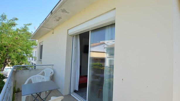 A balcony with a table and chairs, a sliding glass door, and a view of a neighboring building. - Apartment with parking - 6 guests (Le Grau-du-Roi)