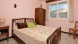 A bedroom with a bed, a nightstand, a wooden wardrobe, a window with curtains, and a chair. - Apartment with parking - 6 guests (Le Grau-du-Roi)