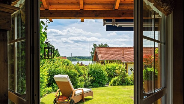 An open door leading to a garden with a lawn, a wicker chair, and a view of a house with a red roof. - Kathi Vacation Home on Lake Jabelschen with a Garden, Terrace, and Fireplace (Jabel)