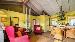 A living room with a red sofa, a red armchair, a dining table with wicker chairs, a chandelier, and a kitchen area with yellow walls and wooden beams. - Kathi Vacation Home on Lake Jabelschen with a Garden, Terrace, and Fireplace (Jabel)