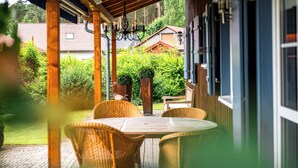 A covered patio with wicker furniture, a wooden table, and a view of a garden and houses in the background. - Kathi Vacation Home on Lake Jabelschen with a Garden, Terrace, and Fireplace (Jabel)