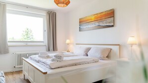 Ein ordentlich bezogenes Bett mit weißen Leinen, eine Nachttischlampe und ein Gemälde mit einem Sonnenuntergang über dem Meer an der Wand.