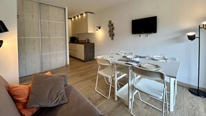 Un saló modern amb un sofà, taula de menjador i cadires, una cuina al fons i un televisor fixat a la paret.