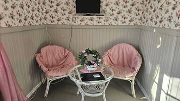 Quarto com papel de parede floral, televisão na parede, duas cadeiras com almofadas, uma mesa pequena com um laptop e flores, e uma janela com luz solar.
