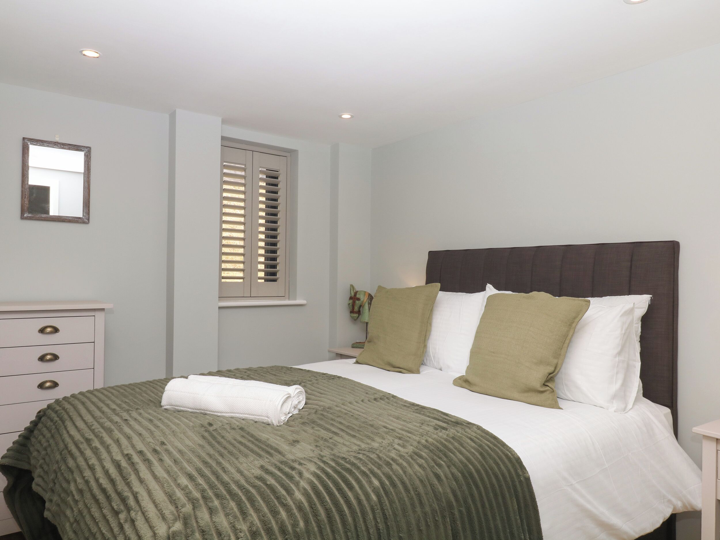 Ein Schlafzimmer mit Bett, Schminktisch, Fenster mit Jalousien und einem Spiegel an der Wand.