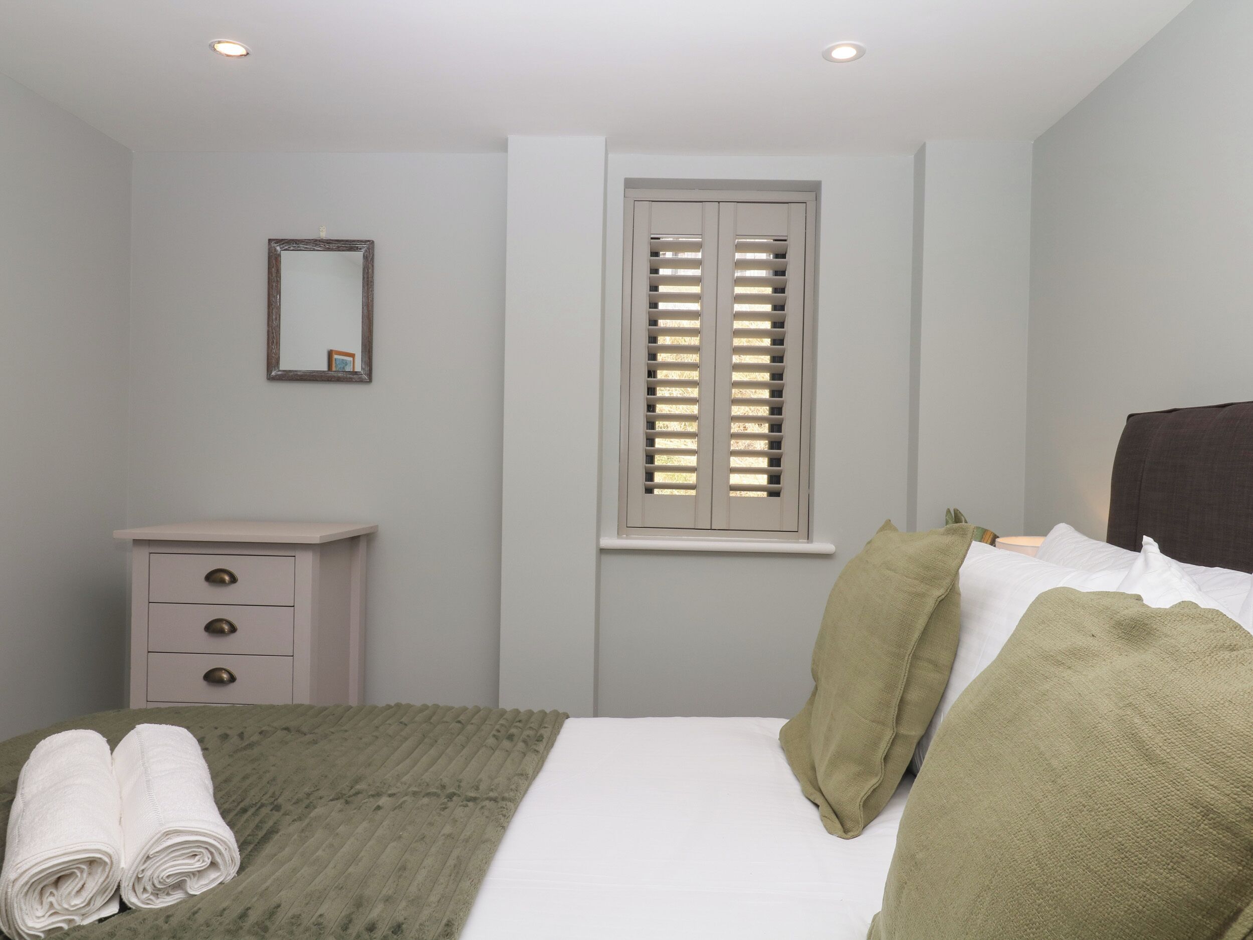 A bedroom with a bed, a bedside table, a mirror, and a window with shutters.