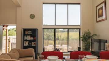 A dining area with a table set for two, a bookshelf, a clock, and a large window with a view of the outdoors.