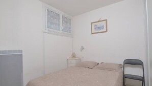 A bedroom with a bed, a chair, a radiator, a window with a curtain, a bedside table with a lamp, and a framed picture on the wall.