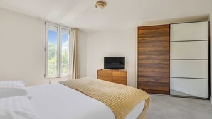 Un dormitorio con cama, televisor sobre un aparador de madera, armario corredero y ventana con cortinas.