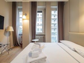 A hotel room with a bed, a small round table, a lamp, and a view of buildings through large windows. - Freguglia G - Modern Studio With Balcony Close to Duomo (Milan)