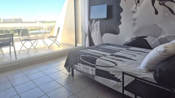A bedroom with a large window, a bed with a patterned bedspread, a mounted TV, and a view of the outside balcony. - Comfortable studio with air conditioning - Cap d'Agde (Cap d'Agde)