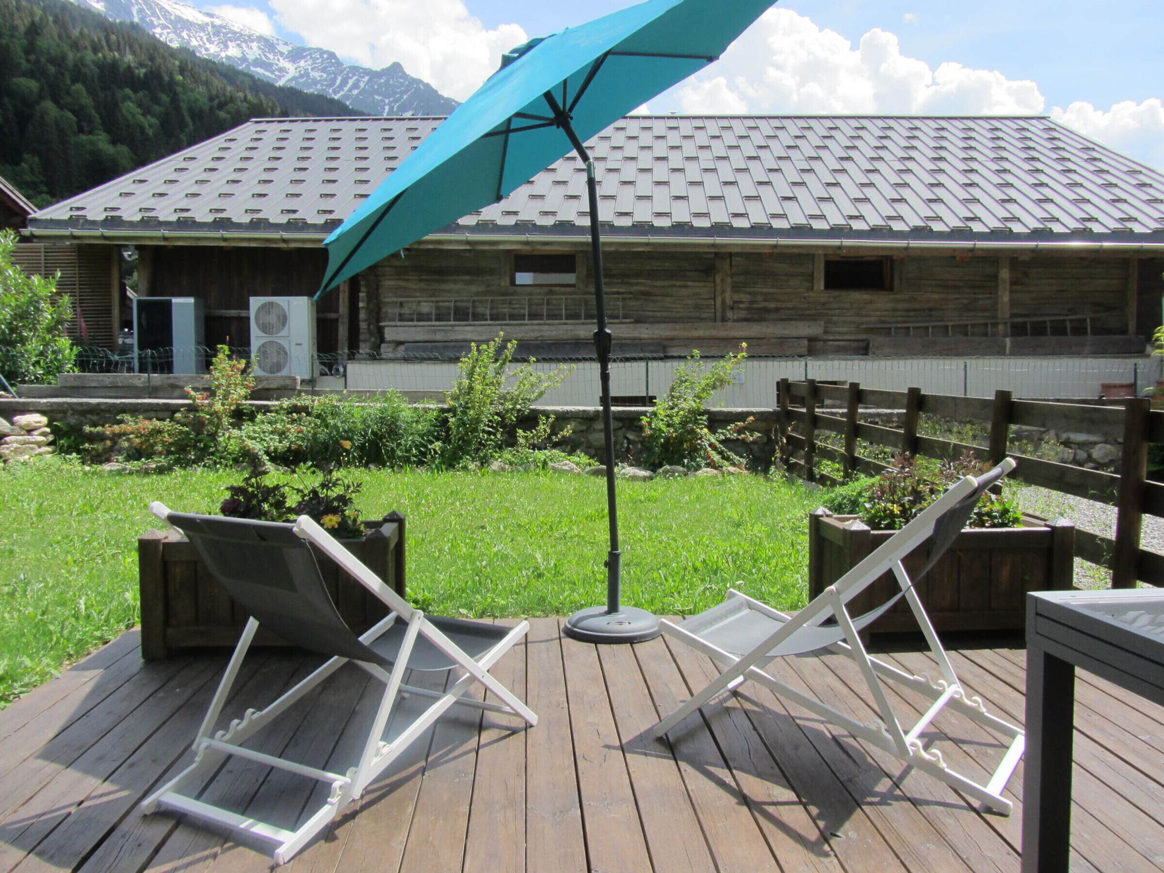 A wooden deck with two lounge chairs and a table, a blue umbrella, and a building with a sloped roof in the background.
