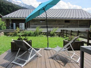A wooden deck with two lounge chairs and a table, a blue umbrella, and a building with a sloped roof in the background. - Spacious apartment, panoramic view - 6 guests (Les Contamines-Montjoie)