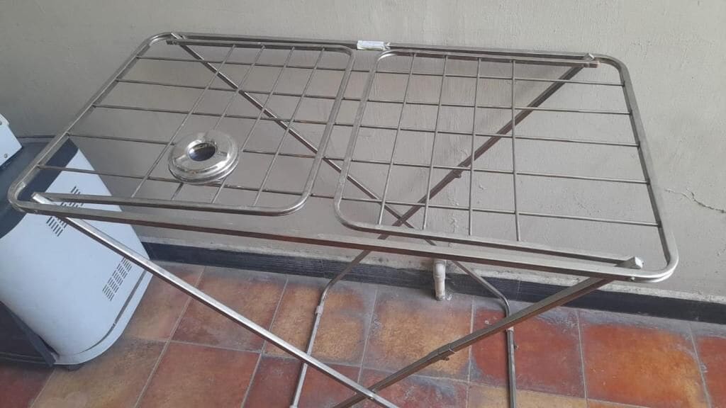 A metal drying rack with a circular base and a grid top, placed on a tiled floor against a wall.