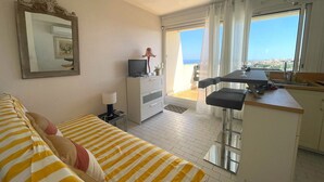 Ett rum med en soffa, en tv-apparat på ett skåp, ett litet bord med en stol och utsikt över havet från en balkong.