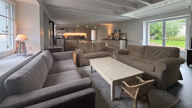 Ein großzügiger Wohnbereich mit einer Couch, einem Couchtisch und einem Korbstuhl. Im Hintergrund ist eine Küche zu sehen.