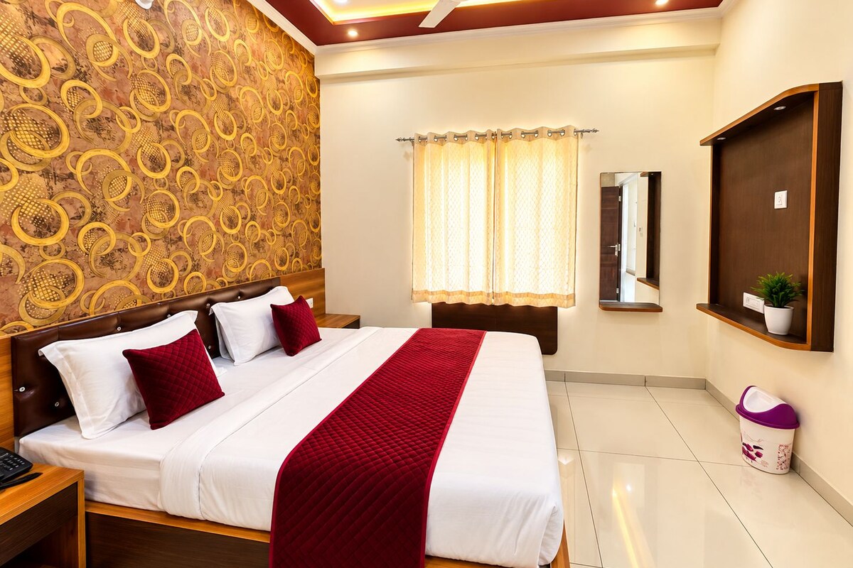 A hotel room with a large bed, a ceiling fan, a window with curtains, a mirror, and a small plant on a shelf.